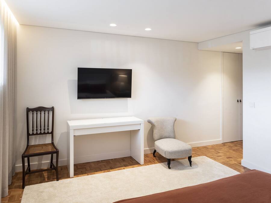 A modern bedroom with a white desk, a television mounted on the wall, a wooden chair, a brown ottoman, and a large bed with a brown bedspread.