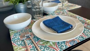 Une table à manger dressée avec un bol blanc, une serviette bleue, un verre à vin et une fourchette, le tout posé sur une nappe à motifs.