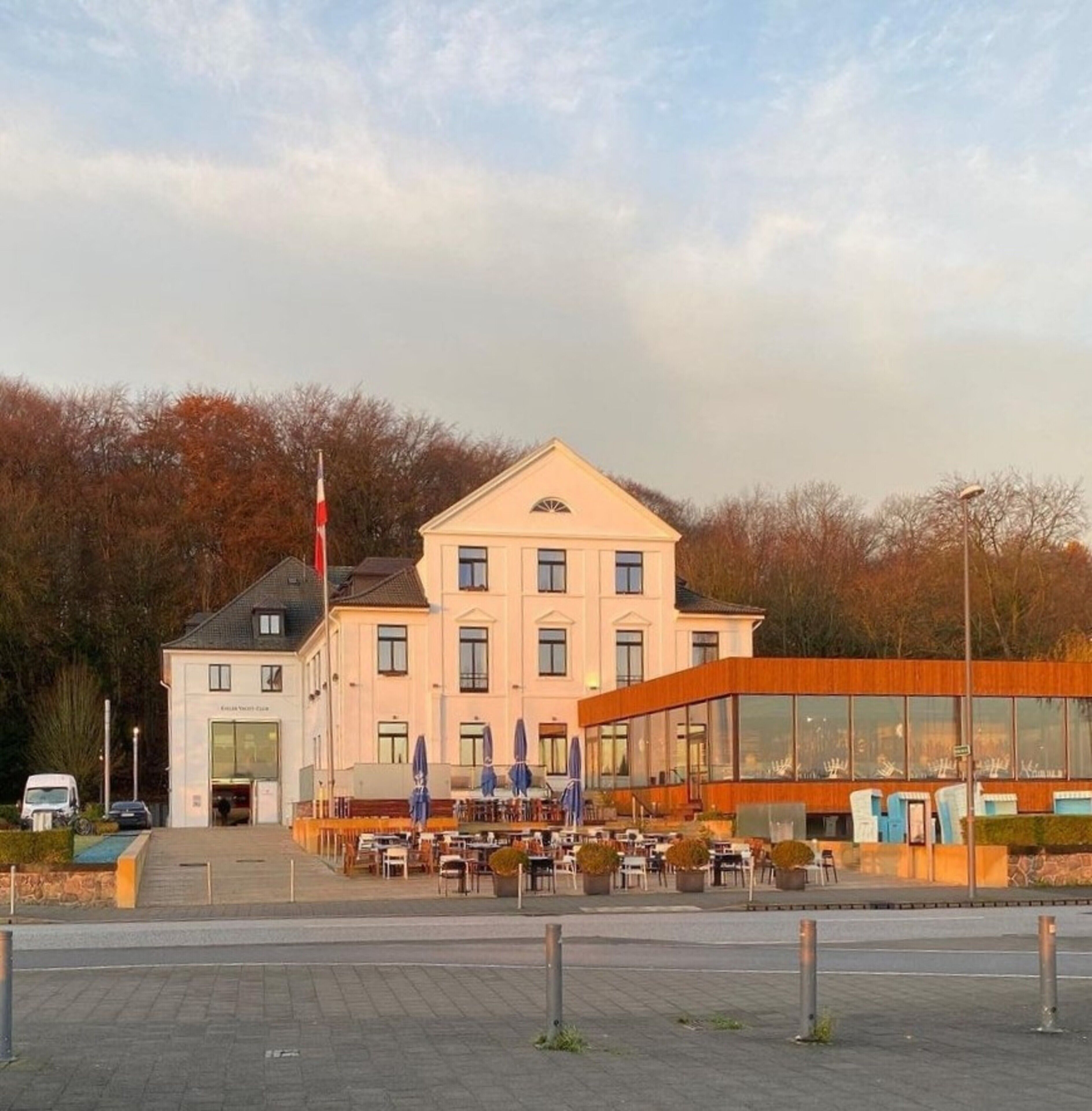 Un grand bâtiment doté d’une entrée centrale, d’un mât où flotte un drapeau et d’un espace aménagé avec des tables et des chaises.