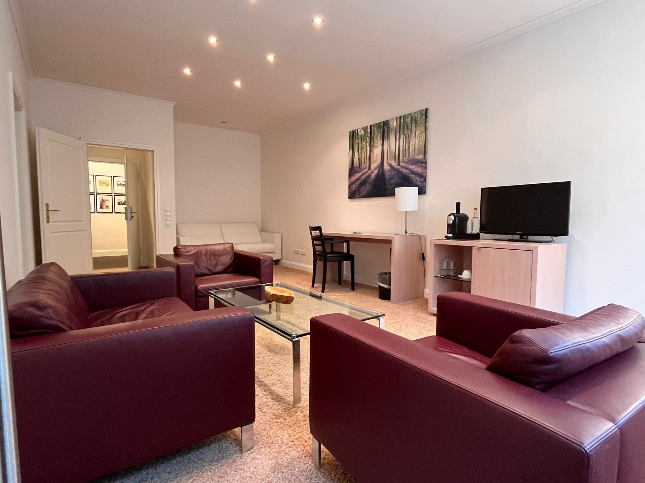 A modern living room with a glass coffee table, two leather sofas, a dining area with a chair, a TV on a cabinet, and a painting on the wall.