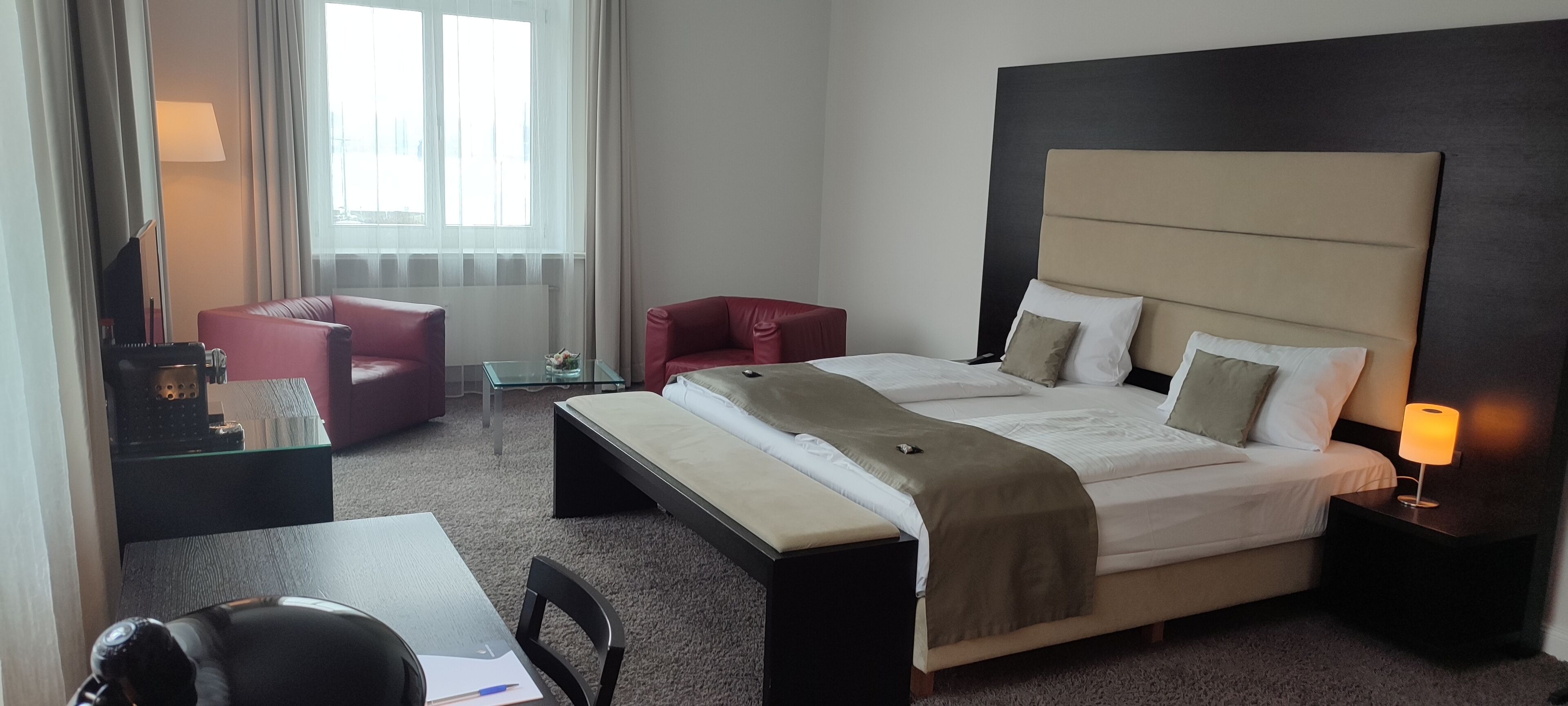 Una habitación de hotel con una cama grande, un escritorio con una silla, un sillón rojo y una mesita de noche con una lámpara.
