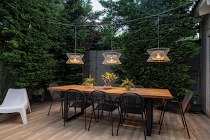 Una mesa de madera para comer con seis sillas, rodeada de plantas en macetas y un muro de piedra.