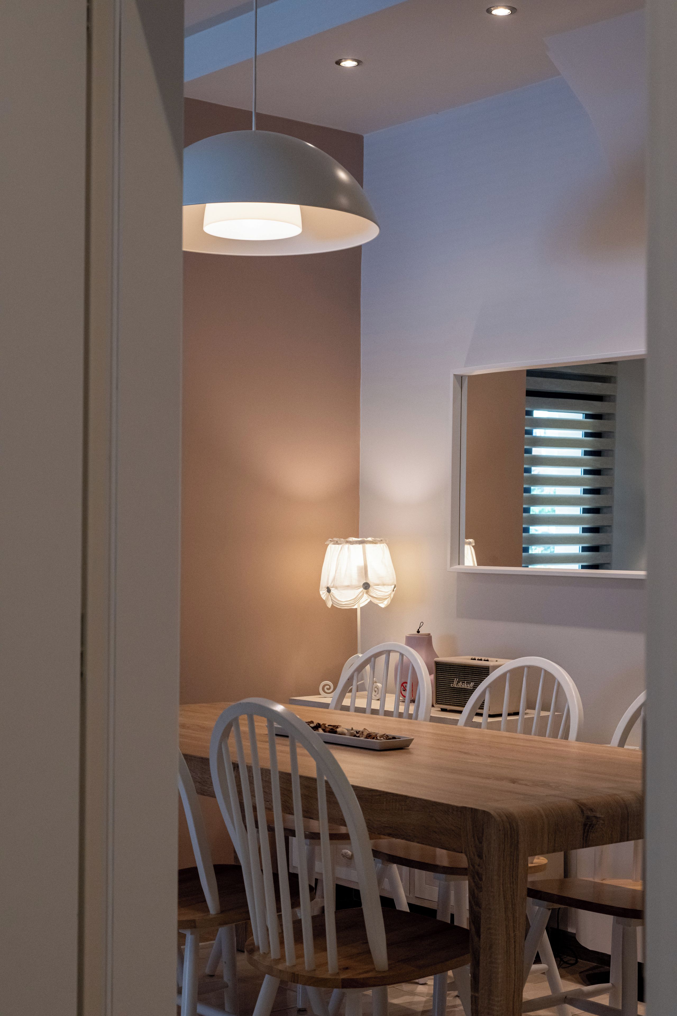 Un coin repas avec une table en bois, des chaises blanches, des luminaires suspendus, une lampe de table et une fenêtre avec des stores.