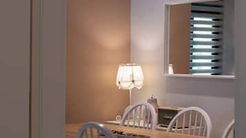 Un coin repas avec une table en bois, des chaises blanches, des luminaires suspendus, une lampe de table et une fenêtre avec des stores.