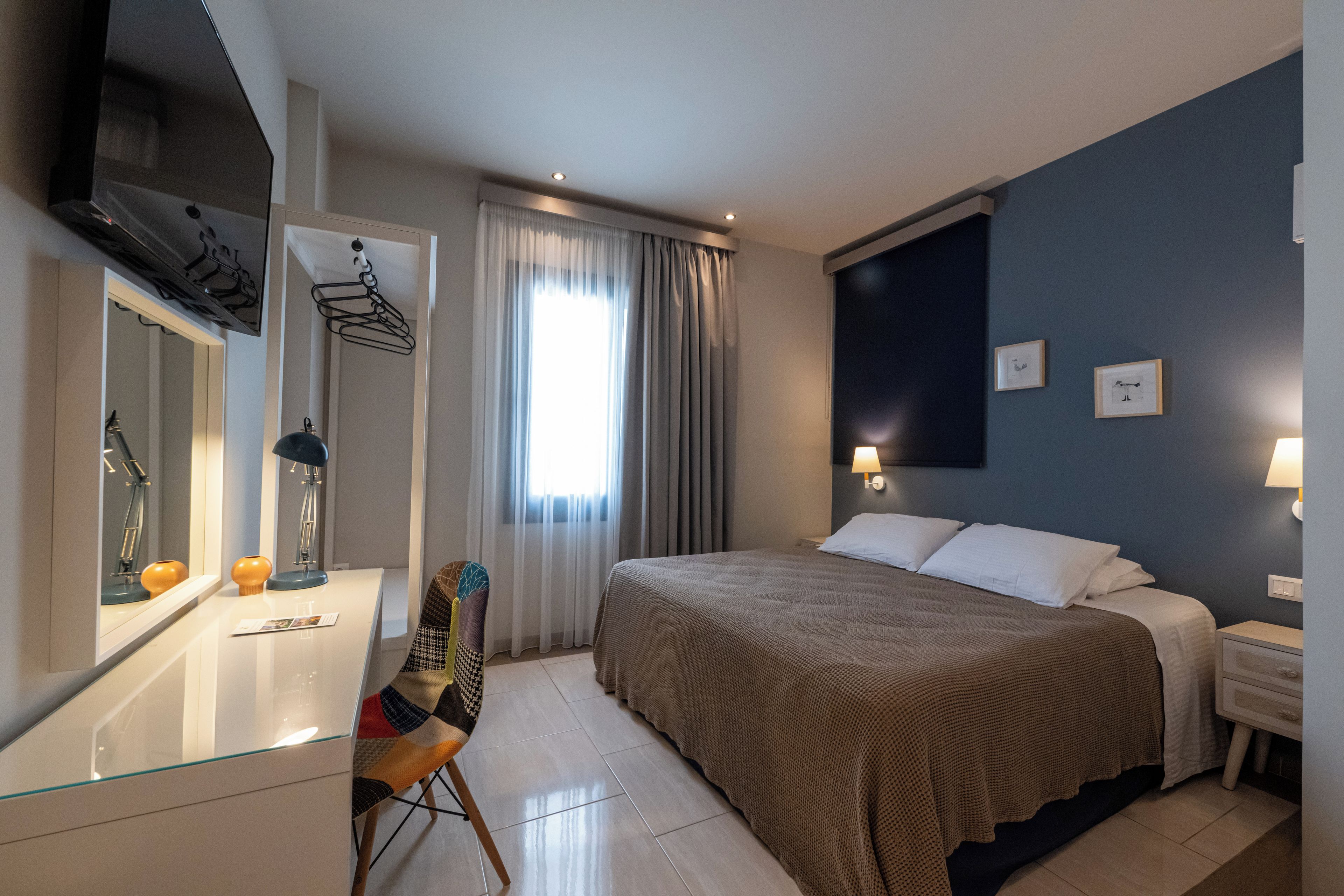 A hotel room with a bed, a desk with a mirror, a chair, a TV, and a window with curtains.