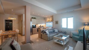 A modern living room with a sofa, coffee table, and TV. A kitchen with cabinets and a dining area with chairs is visible. - Thalias Quarters - Kalamata Mediterranean Suites & Villas (Kalamata)