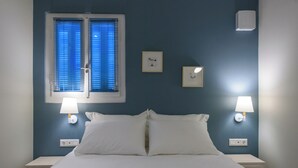 A hotel room with a bed, two bedside lamps, a window with blinds, and wall-mounted light switches. - Thalias Quarters - Kalamata Mediterranean Suites & Villas (Kalamata)