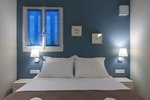A hotel room with a bed, two bedside lamps, a window with blinds, and wall-mounted light switches. - Thalias Quarters - Kalamata Mediterranean Suites & Villas (Kalamata)