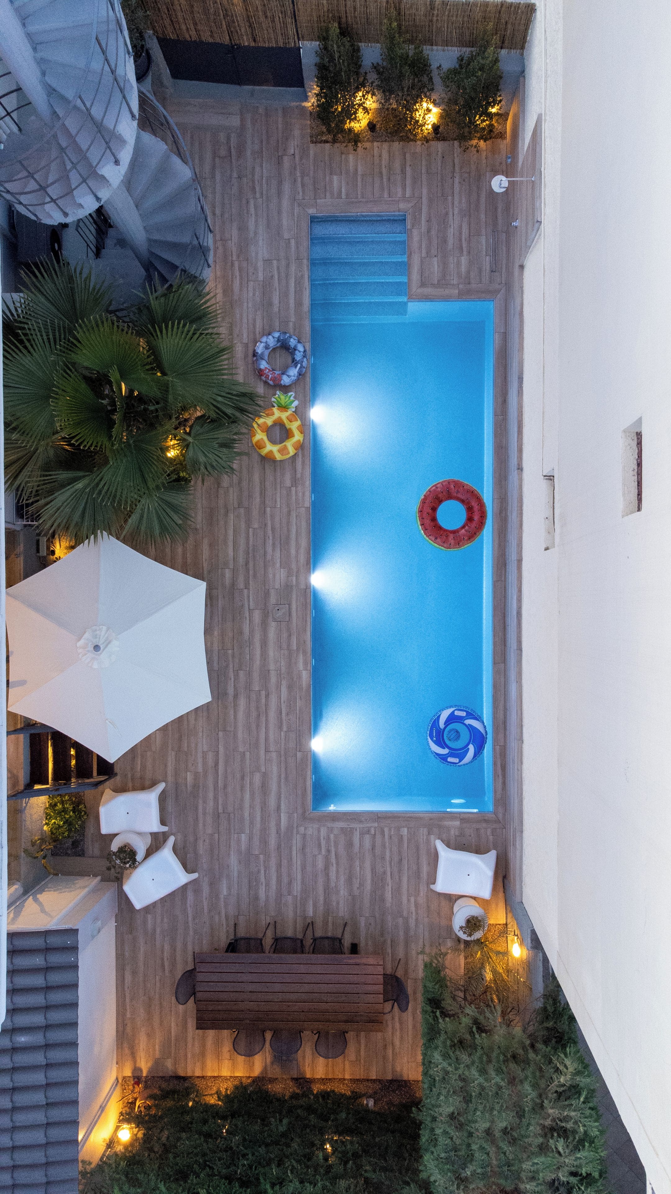 A rooftop pool with a wooden deck, surrounded by lounge chairs and umbrellas, and equipped with a wooden bench and a small table.