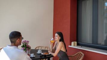 A dining area with a table set for two, featuring a wicker chair, a vase with flowers, and a glass of orange juice.