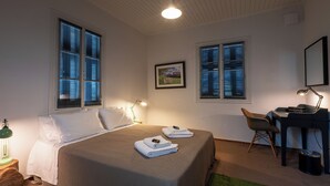 Ein Schlafzimmer mit einem Bett, zwei Nachttischlampen, einem Schreibtisch mit Stuhl und zwei Fenstern mit Jalousien.