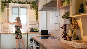 A kitchen with a woman standing by the counter, a washing machine, and various kitchen appliances. - Greenhouse - Kalamata Mediterranean Suites & Villas (Kalamata)
