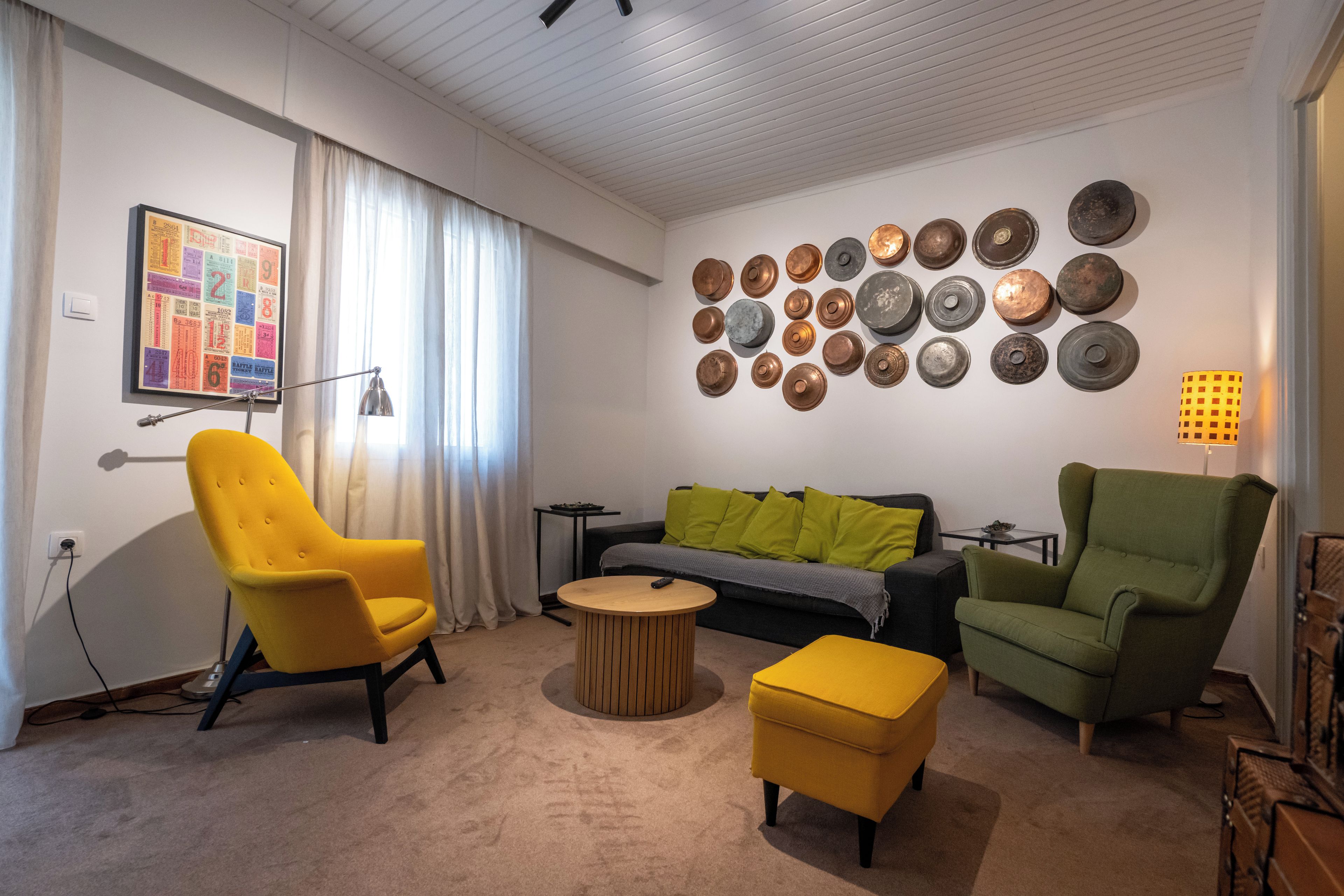Una sala de estar con un sillón amarillo, un sillón verde, una mesa de centro redonda y un otoman amarillo. Se observa una pared con múltiples platos decorativos y una fotografía enmarcada.