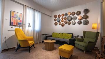 Una sala de estar con un sillón amarillo, un sillón verde, una mesa de centro redonda y un otoman amarillo. Se observa una pared con múltiples platos decorativos y una fotografía enmarcada.