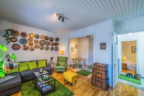 A living room with a sofa, armchair, coffee table, and a dining area with a table and chairs. There are wall decorations and a ceiling fan. - Greenhouse - Kalamata Mediterranean Suites & Villas (Kalamata)