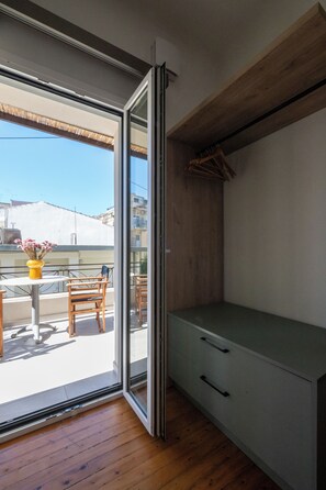 A room with a sliding glass door leading to a balcony with wooden furniture and a yellow vase with flowers.