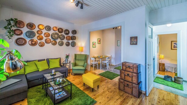 Ein Wohnzimmer mit Sofa, Sessel, Couchtisch und einem Essbereich mit Tisch und Stühlen. An den Wänden befinden sich Dekorationen und es gibt einen Deckenventilator.