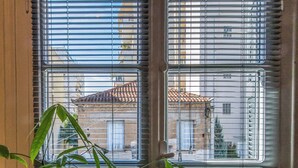 A window with metal blinds, a view of parked cars, and a building with a tiled roof in the background. - Greenhouse - Kalamata Mediterranean Suites & Villas (Kalamata)