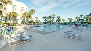 Zona de piscina con sillas de descanso y palmeras, rodeada por un edificio con amplias ventanas.