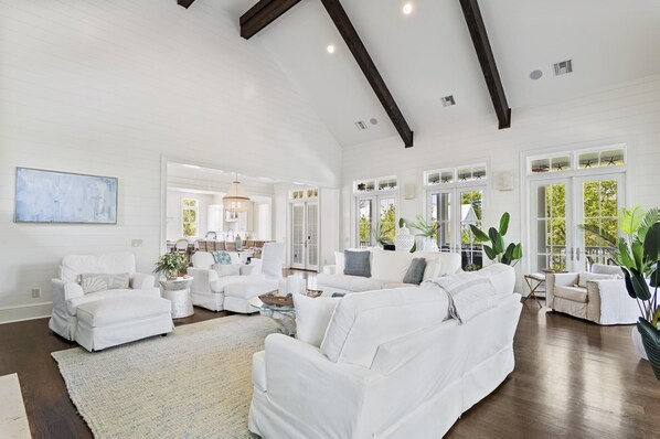 A spacious living room with white furniture, large windows, and a high ceiling with exposed beams.