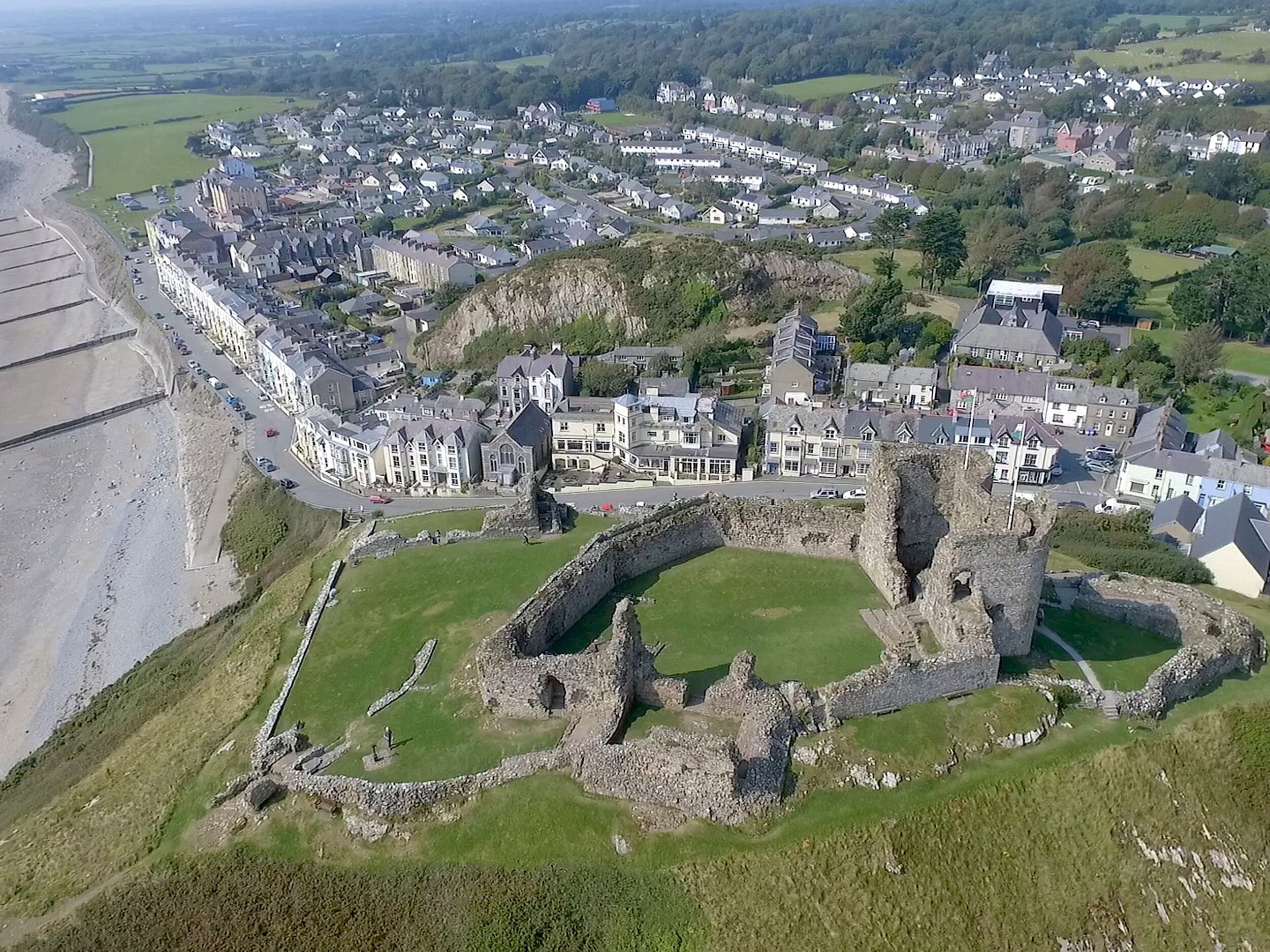 An aerial view of a coastal town with historic stone ruins, residential buildings, and a beach.