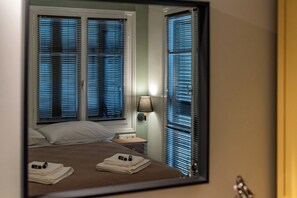 A hotel room with a bed, a nightstand, a lamp, and a window with blinds. - Tinas Home - Kalamata Mediterranean Suites & Villas (Kalamata)