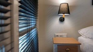 A bedroom with a bedside table, a lamp, and a window with blinds. - Tinas Home - Kalamata Mediterranean Suites & Villas (Kalamata)
