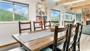 A dining area with a wooden table and chairs, large windows with a mountain view, and a stone wall. - Trails End Vacation Home At Windcliff 3 Bedroom Home by RedAwning (Estes Park)