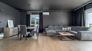 A modern living room with a grey sectional sofa, a wooden dining table with chairs, and a large window with curtains. - Villa Marisa with 51sqm heated, private pool, 5 bedrooms, 4 bathrooms, summer kitchen, views of the Adriatic sea and town Split (Kastel Kambelovac)
