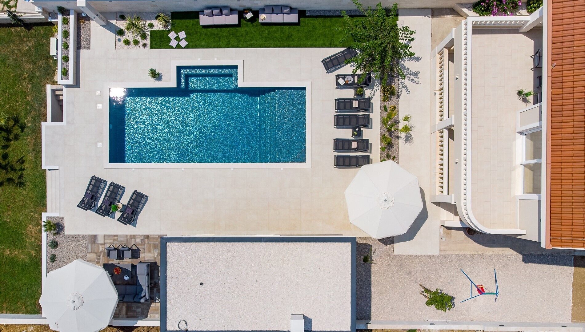 Vista aerea di un hotel moderno con piscina centrale, lettini prendisole e un’area salotto coperta.