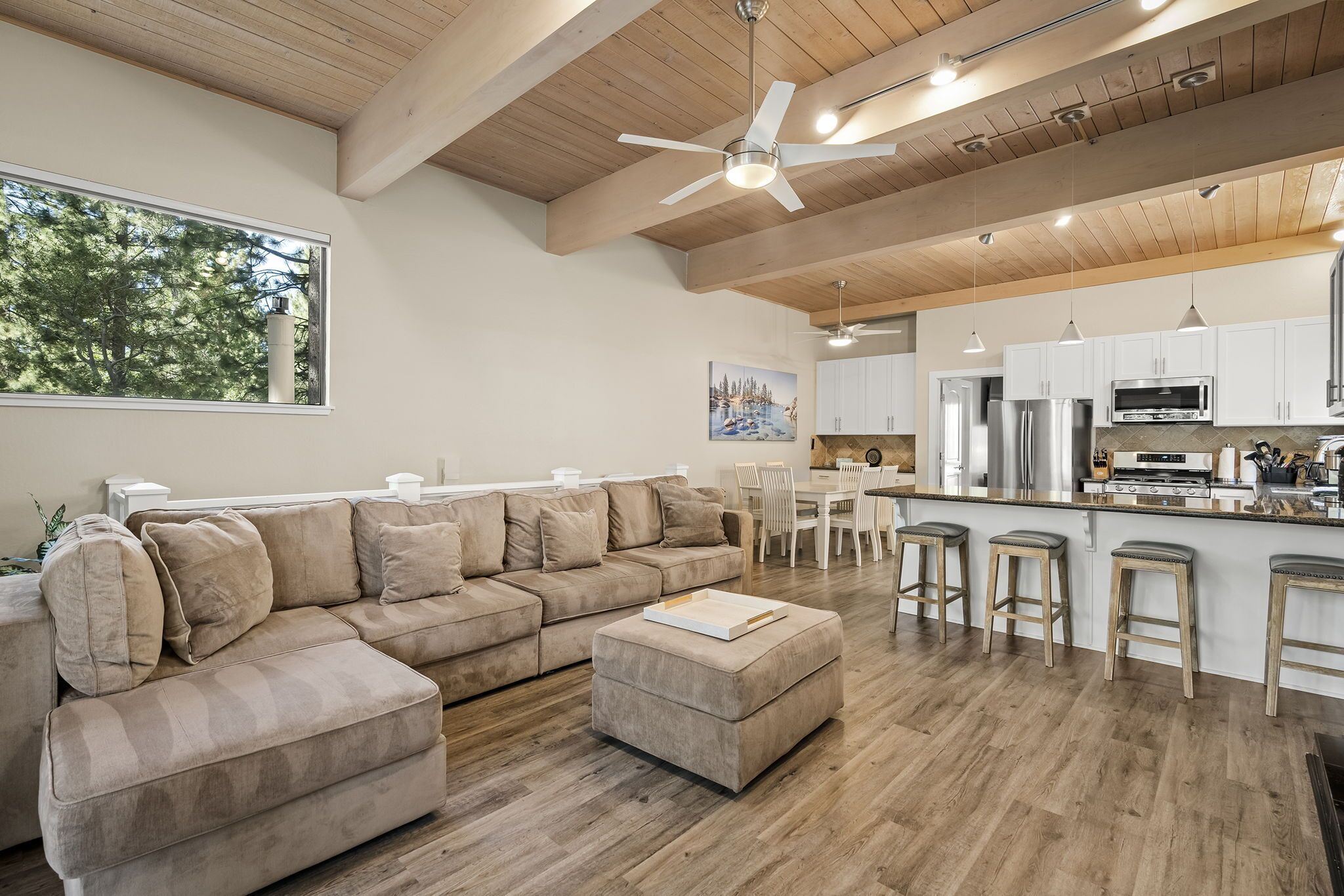 A modern living room with a beige sectional sofa, a wooden ceiling, and a kitchen area with white cabinets and stainless steel appliances.