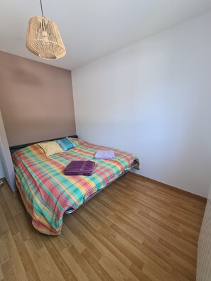 Ein kleines Zimmer mit einem Einzelbett, Holzboden, bunter Karo-Bettdecke und einer gewobenen Pendelleuchte.