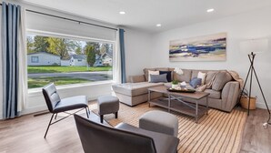 A modern living room with a sectional sofa, a coffee table, and a view of a suburban neighborhood. - CDA ID. Sanders Beach! Best Location! Newly Remodeled Addition! Pet Friendly. (Coeur d'Alene)
