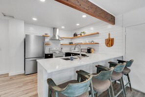 A modern kitchen with white cabinetry, a stainless steel refrigerator, a central island with bar stools, and wooden beams on the ceiling.