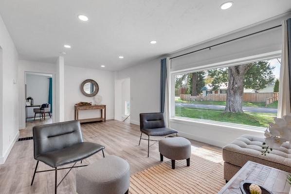A modern living room with a large window, a wooden console table, two leather chairs, a grey ottoman, and a sofa.