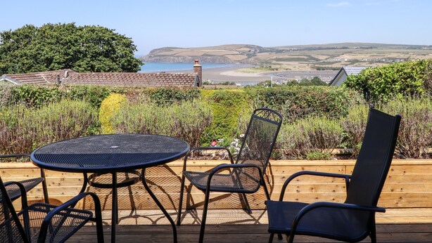 Une terrasse dotée d’une table ronde et de chaises, offrant une vue sur un paysage côtier avec des collines et une plage.
