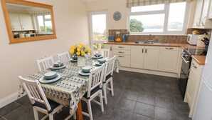 Une cuisine avec une table à manger dressée pour quatre personnes, une horloge au mur et une vue sur l’extérieur par la fenêtre.