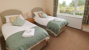 Une chambre double avec deux lits, chacun recouvert d’une couverture verte et agrémenté d’une serviette pliée. La chambre est dotée d’une fenêtre avec des rideaux, d’un radiateur et d’une table de chevet avec une lampe.