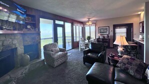 A living room with a stone fireplace, a large window with a view, a dining area, and a seating area with a lamp. - Beautiful Sugar Mountain Condo with Long-Range Views (Sugar Mountain)