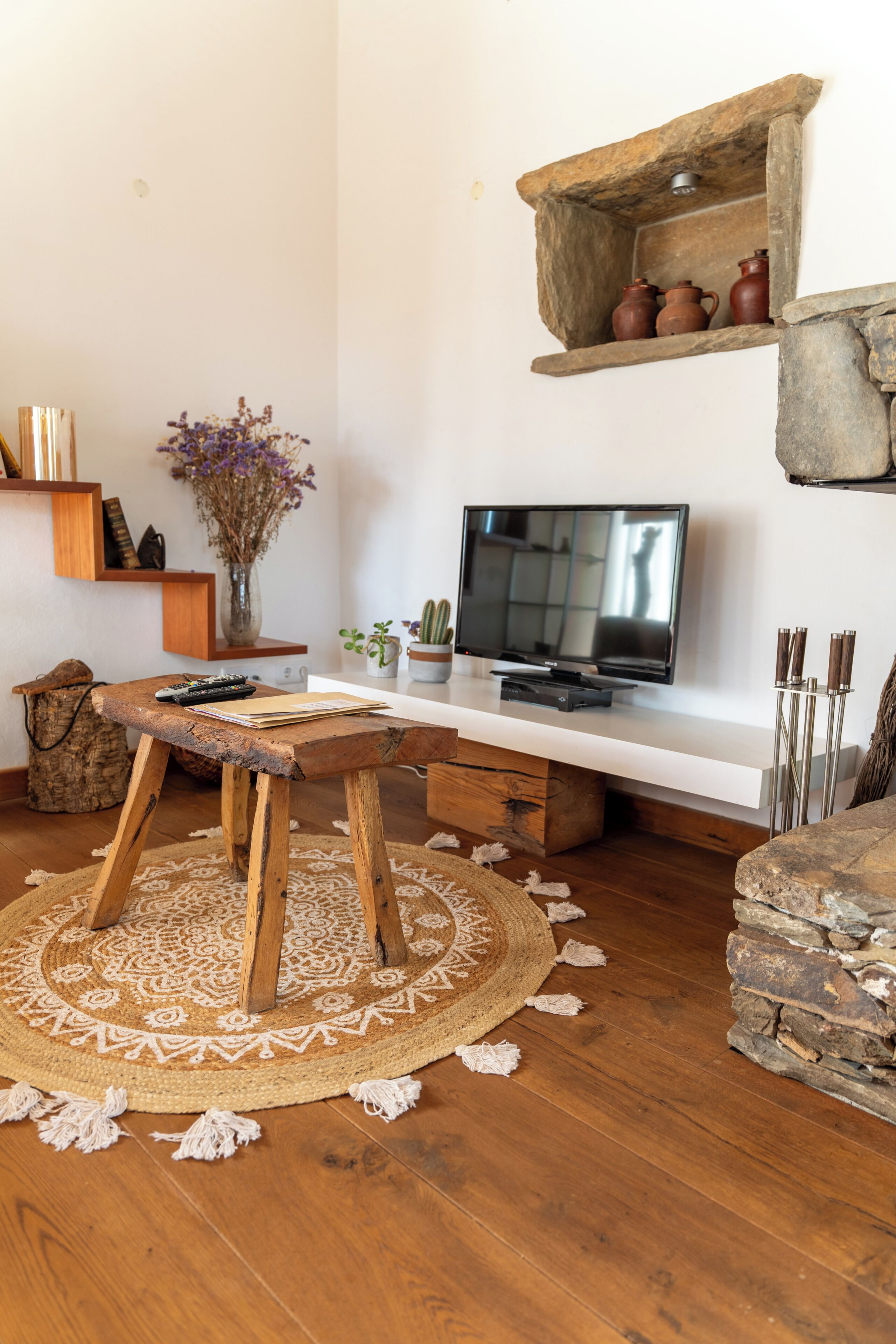 Una mesa de madera con un control remoto, un televisor sobre un mueble, un tapete tejido y una estantería de piedra con cerámica.