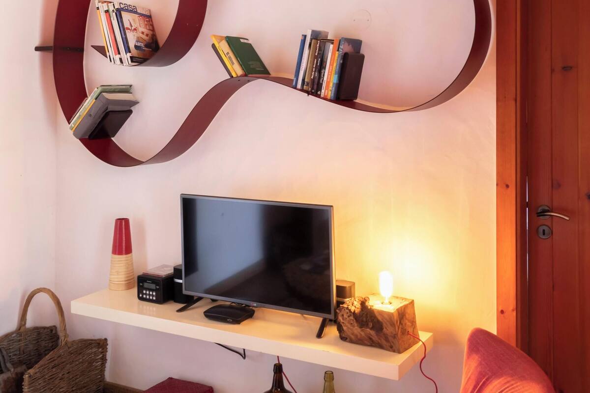 Ein Wohnzimmer mit Fernseher, Bücherregalen, einem roten Sessel, einem Holztisch und einem Korb.