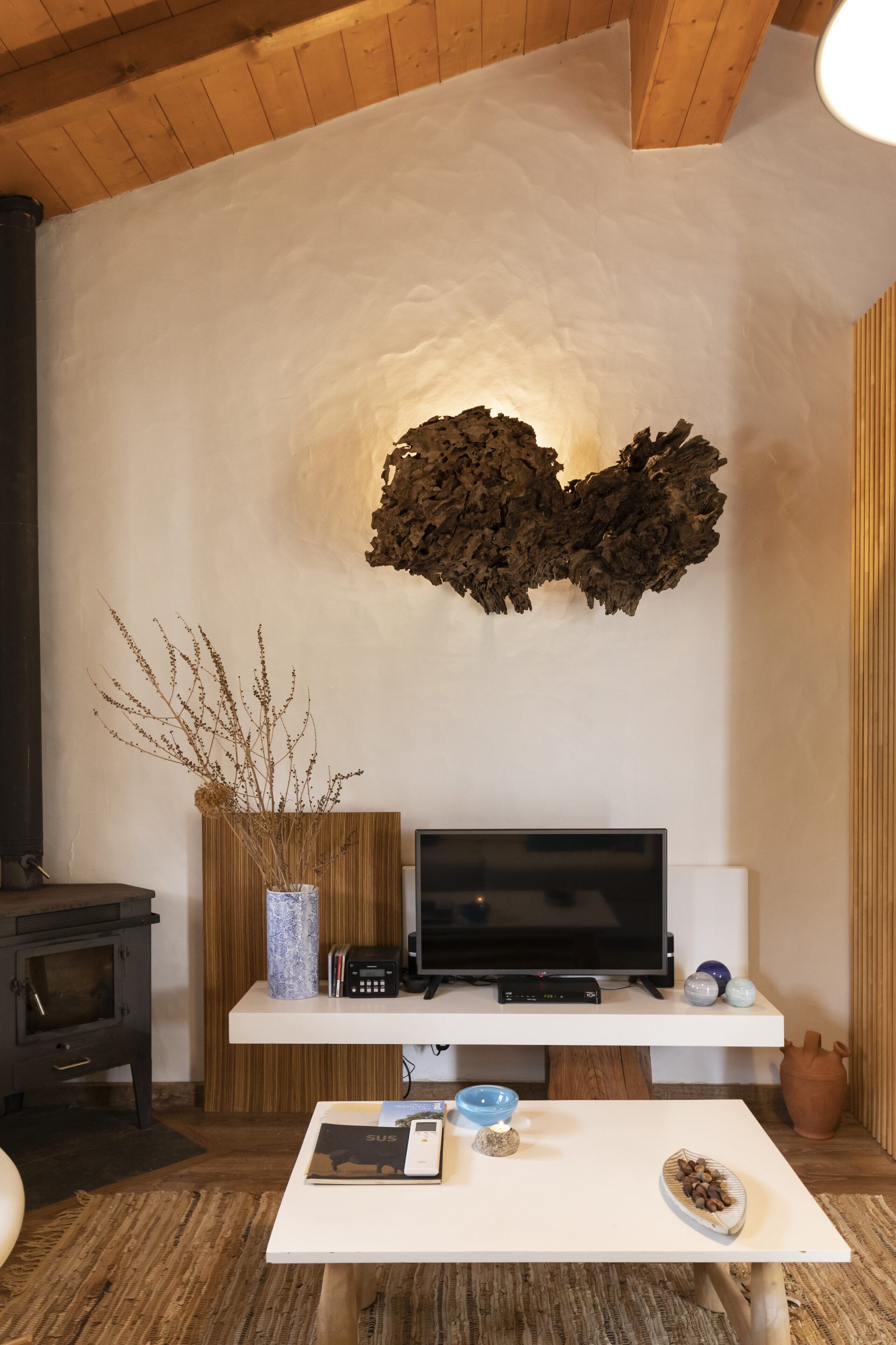 Un soggiorno moderno con una TV a schermo piatto su un mobile bianco, una decorazione a parete in legno, un vaso con piante essiccate, una ciotola e un tavolino con un telecomando.