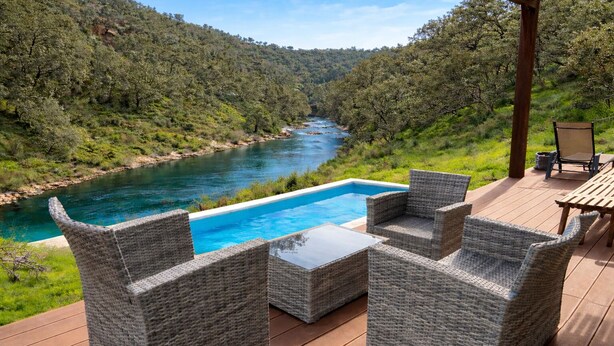 A deck with wicker furniture overlooking a river and forested hills.