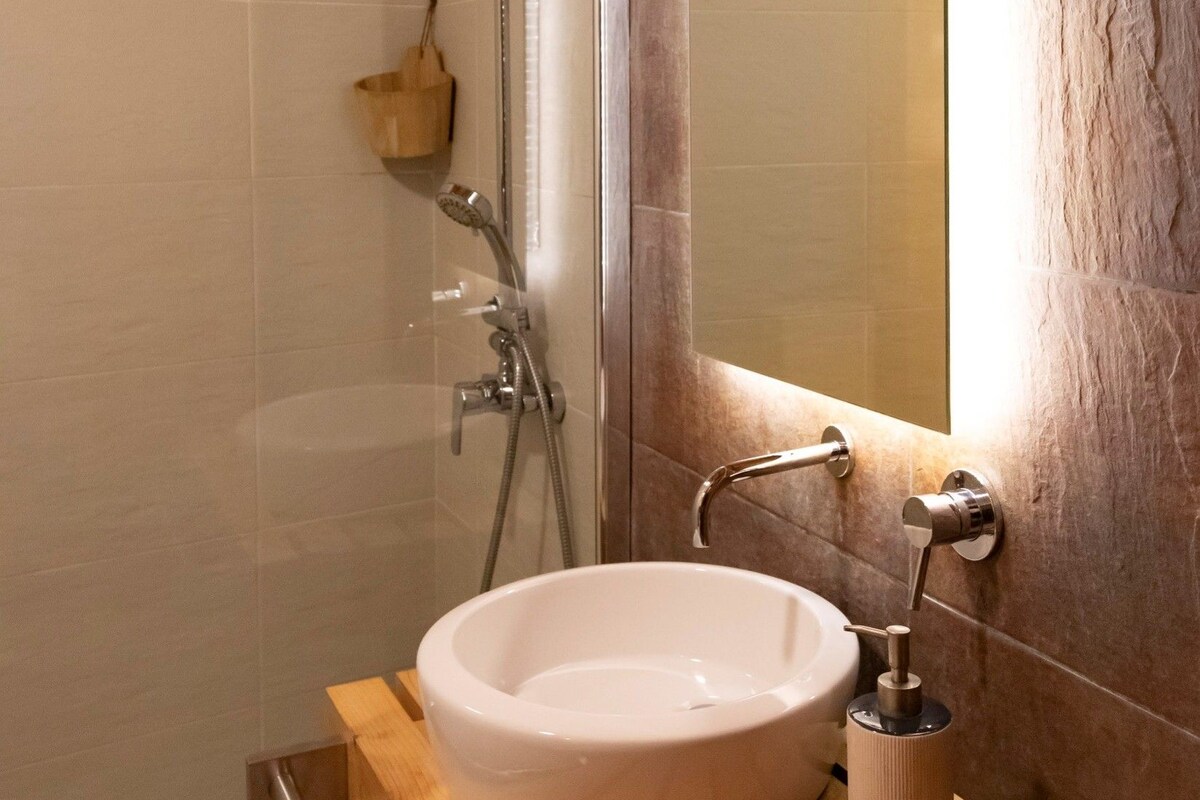 Une salle de bain avec un lavabo rond, une douchette à main, un meuble-lavabo en bois et un miroir avec un cadre en bois.