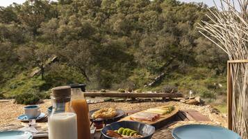 A breakfast setup with a bottle of milk, a bowl of fruit, and a plate of food on a table with a view of a forested hillside.