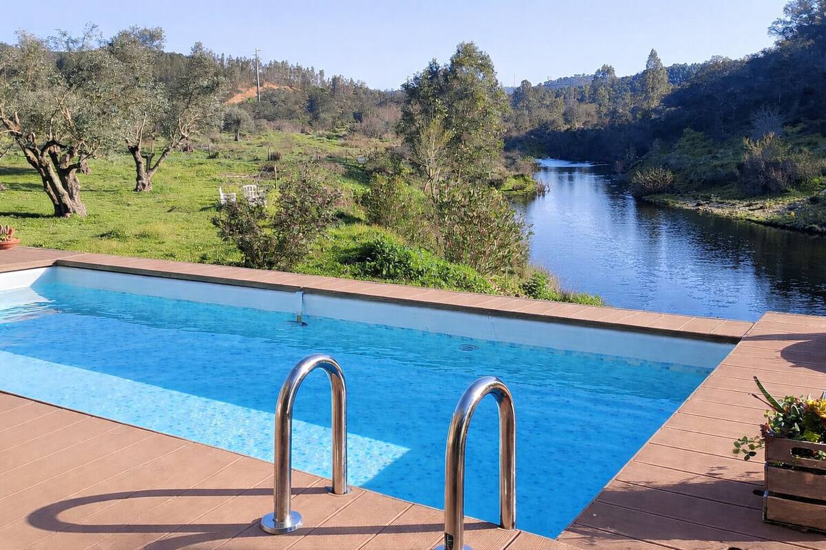 Ein rechteckiger Pool mit Metallleitern, umgeben von einer Holzterrasse, mit Blick auf einen Fluss und Grünflächen.