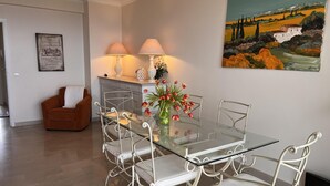 Een eetgedeelte met een tafel met glazen blad, omringd door witte metalen stoelen, een bloemenvaas en twee lampen op een kast.