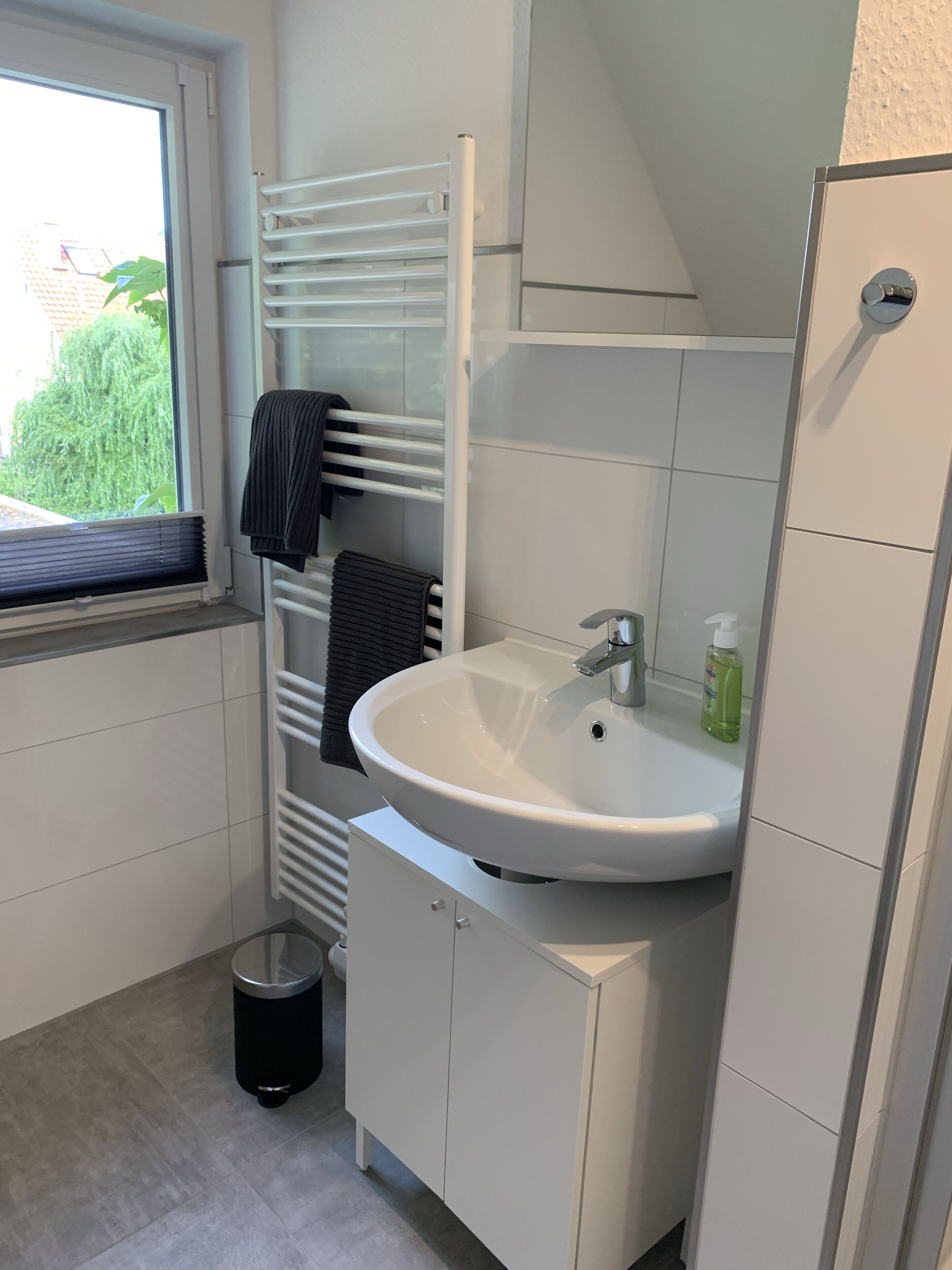 Ein kleines, modernes Badezimmer mit einem weißen Waschbecken, einem Handtuchhalter, einem Fenster und einer Mülleimer.