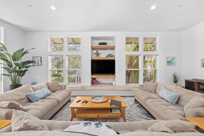 Una sala de estar moderna con un conjunto de sofás, una mesa de centro, un televisor de pantalla plana y ventanas amplias con vistas a zonas verdes.