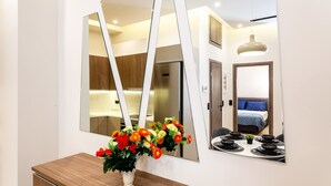 Un espejo con un mueble de madera debajo, un jarrón con flores y el reflejo de una habitación con una cama y una lámpara colgante.